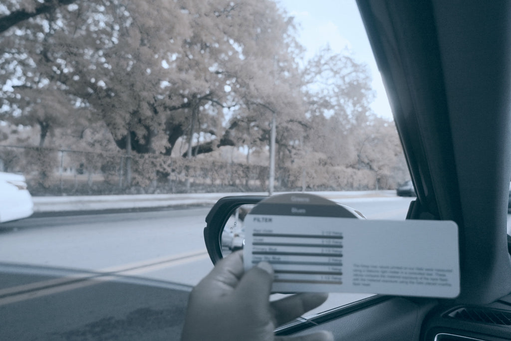 Bleach Bypass profile added after photo example - A hand holding a white rectangular card in front of a car's side mirror and window, with a blurred road and trees visible outside.