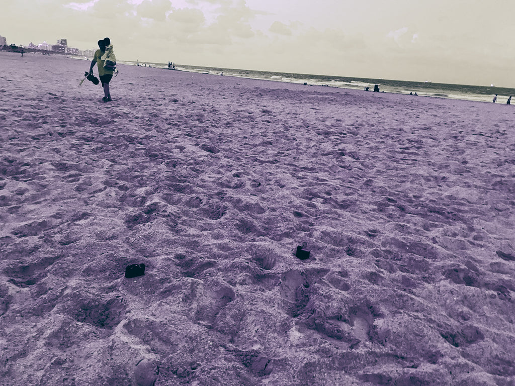 A wide sandy beach scene with a person carrying a large black case walking near the shoreline under an overcast sky. The image has a strong monochrome purple cast.